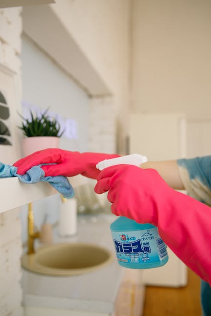 Two professional cleaning staff members from Cleaner Clapham are standing in a well-lit, white room, dressed in beige cleaning uniforms with blue badges. They are smiling at the camera, each holding cleaning tools: one with a red vacuum cleaner and the other with a green mop. The surfaces around them are clean, reflecting thorough surface cleaning and sanitisation, with no visible dust or dirt. Their poised stance suggests readiness for domestic cleaning tasks, emphasizing quality hygiene and deep cleaning services offered by Cleaner Clapham for residential or commercial property maintenance.