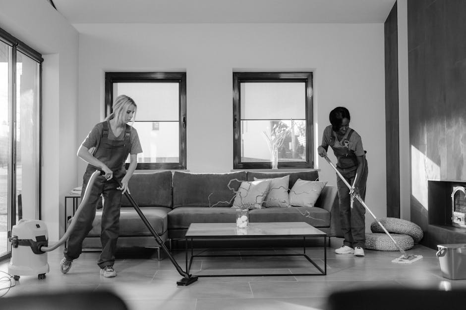 Two professional cleaners from Cleaner Clapham are engaged in surface cleaning within a modern living room. One cleaner, on the left, is vacuuming the wooden floor using a cylinder vacuum cleaner, whi